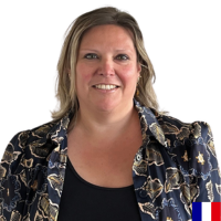 Portrait of a smiling woman with blonde hair wearing a patterned blazer on a white background, with a small French flag in the bottom-right corner.